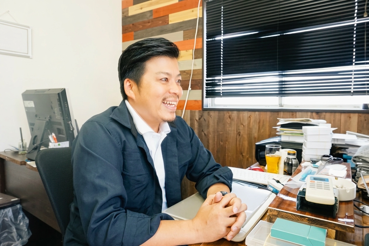 作業着姿で事務所のデスクに座り笑顔で打ち合わせをする工事事業者の男性